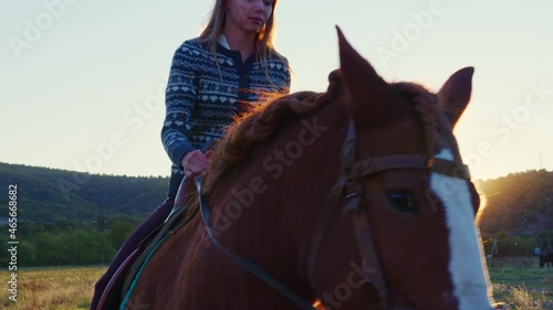 Beautiful girl rides horse in pasture surrounded by forests, hills. Young woman goes on horseback in countryside. Light and shadow. Sun shining above mountain. Horsewoman is on saddle. Palfrey walks photo