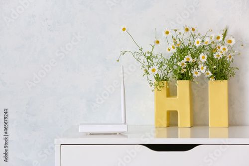 Modern wi-fi router and chamomile flowers on table near light wall