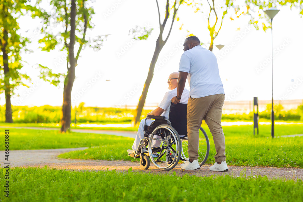 Caregiver and old man in a wheelchair. Professional nurse and patient walking outdoor in the park at sunset. Assistance, rehabilitation and health care. Caregiver and old man in a wheelchair. Professional nurse and patient walking outdoor in the park at sunset. Assistance, rehabilitation and health care.