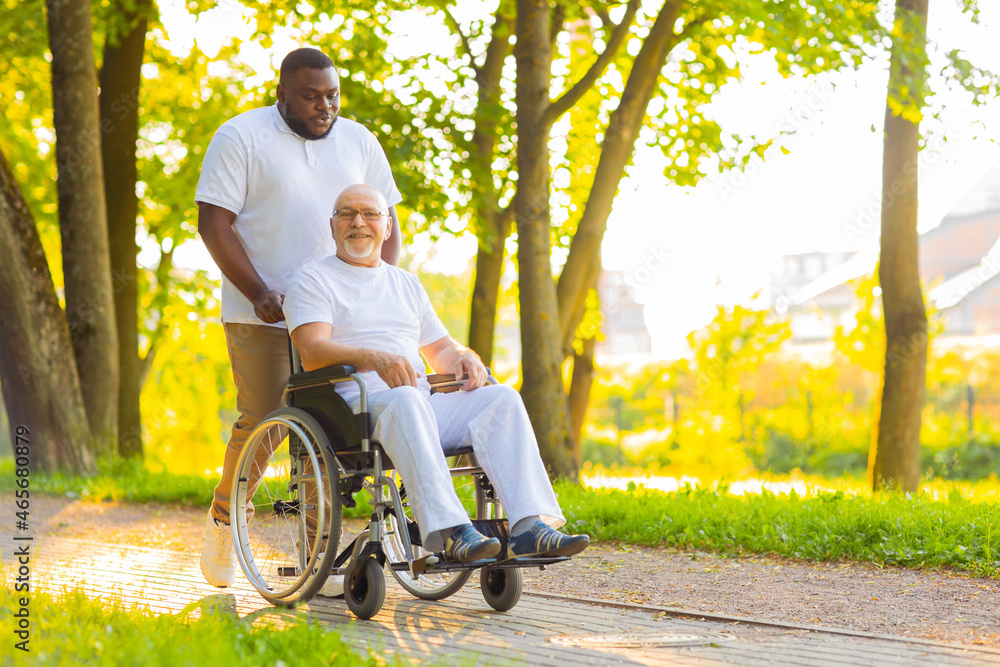Caregiver and old man in a wheelchair. Professional nurse and patient walking outdoor in the park at sunset. Assistance, rehabilitation and health care. Caregiver and old man in a wheelchair. Professional nurse and patient walking outdoor in the park at sunset. Assistance, rehabilitation and health care.