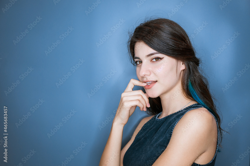 Surprised tender young woman of 20s holding finger at mouth open isolated on blue background studio portrait. With a mysterious pensive expression flirts Surprised tender young woman of 20s holding finger at mouth open isolated on blue background studio portrait. With a mysterious pensive expression flirts