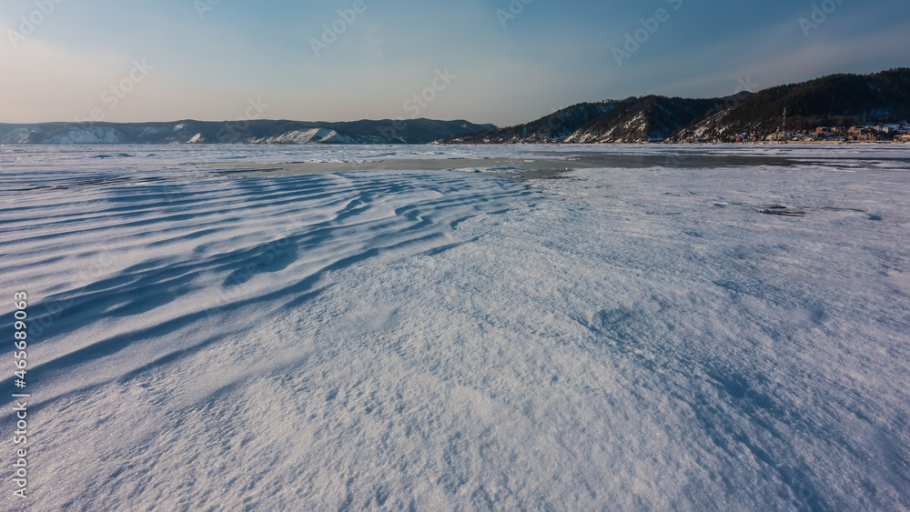 Frozen Lake Baikal. The snow on the surface forms wavy patterns similar to dunes. A mountain range against a blue sky.
