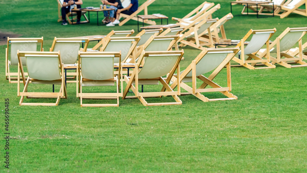 Many empty white deck chairs with tables in lawn is surrounded by shady green grass. Comfortable on outdoor patio chairs in garden. Lawn chairs in the park. Sunbeds in the garden. Selective focus. Many empty white deck chairs with tables in lawn is surrounded by shady green grass. Comfortable on outdoor patio chairs in garden. Lawn chairs in the park. Sunbeds in the garden. Selective focus.