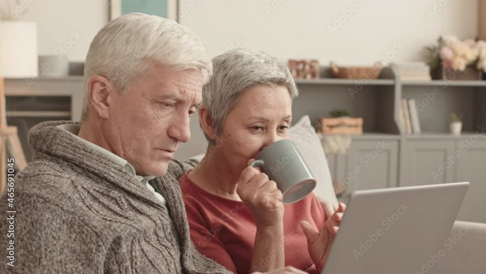 Medium close-up of cheerful senior Caucasian man hugging his Asian wife who holding mug, couple using laptop computer and talking, sitting on sofa at home Medium close-up of cheerful senior Caucasian man hugging his Asian wife who holding mug, couple using laptop computer and talking, sitting on sofa at home