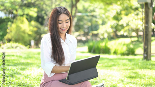 Pleased asian woman sitting in park and using computer tablet.