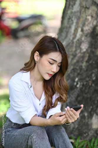 Young asian woman using smart phone while sitting on green grass in park.