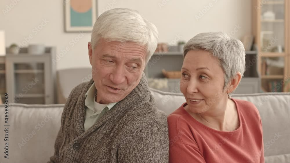 Chest-up side view of happy senior Caucasian man and Asian woman sitting back-to-back on couch in living room, turning heads, looking at each other, talking Chest-up side view of happy senior Caucasian man and Asian woman sitting back-to-back on couch in living room, turning heads, looking at each other, talking