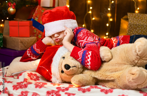 Child girl posing and dreaming in new year or christmas decoration. Holiday lights and gifts, Christmas tree decorated with toys. She's wearing a red sweater and a Santa helper hat