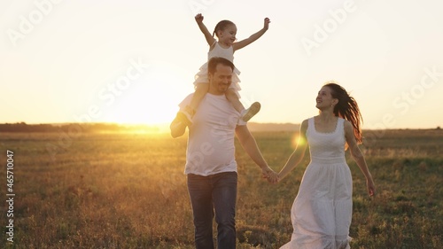 happy family, mother, father and little child, kid sits on shoulders dad, childhood dream of flying, parents with their daughter run at sunset, adult man and woman laugh playing with baby in sunlight