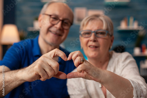 Elder couple creating heart shape sign with hands at home. Married old people in love doing romantic symbol while looking at camera in living room. Husband and wife showing affection figure