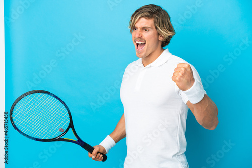 Young blonde man isolated on blue background playing tennis and celebrating a victory