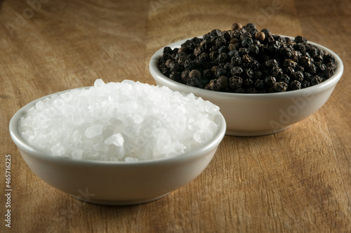Black pepper corns and salt crystals in small white bowls on timber table