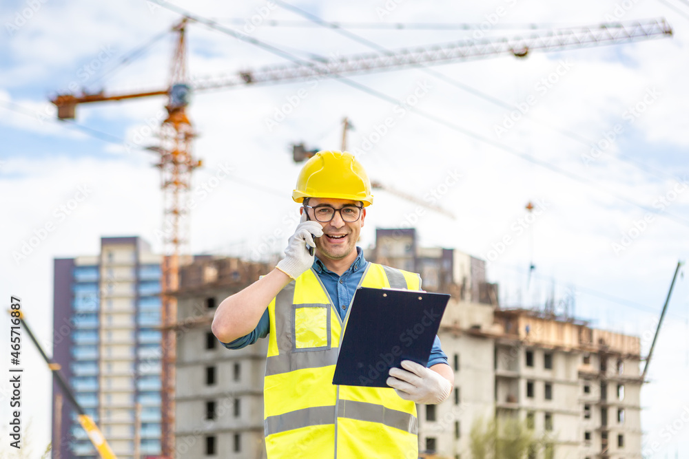 A handsome builder in glasses and a protective helmet stands with documents and talks on the phone against the background of a construction site. A handsome builder in glasses and a protective helmet stands with documents and talks on the phone against the background of a construction site.