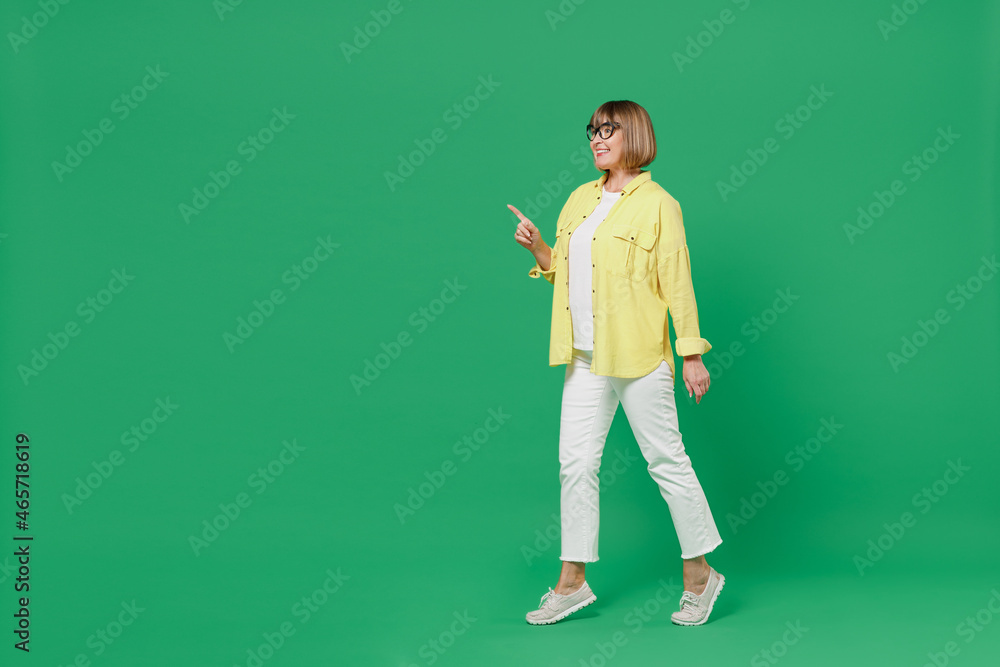 Full body elderly smiling happy woman 50s in glasses yellow shirt point index finger aside on workspace area mock up walk isolated on plain green background studio portrait People lifestyle concept. Full body elderly smiling happy woman 50s in glasses yellow shirt point index finger aside on workspace area mock up walk isolated on plain green background studio portrait People lifestyle concept.