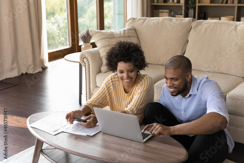 Happy millennial african american family couple analyzing financial paper documents, managing household budget or paying for services in e-banking computer app, enjoying calculating expenses at home.