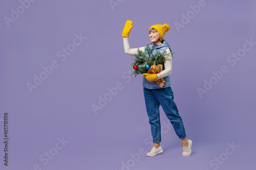 Full size body length mature lady elderly woman 55 years old wears blue waistcoat yellow hat mittens holds spruce branches waving hand isolated on plain pastel light violet background studio portrait.