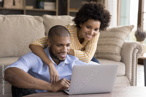 Affectionate young african american woman cuddling shoulders of happy husband, looking at laptop screen, reading email with good news, choosing goods in internet store, web surfing together at home.