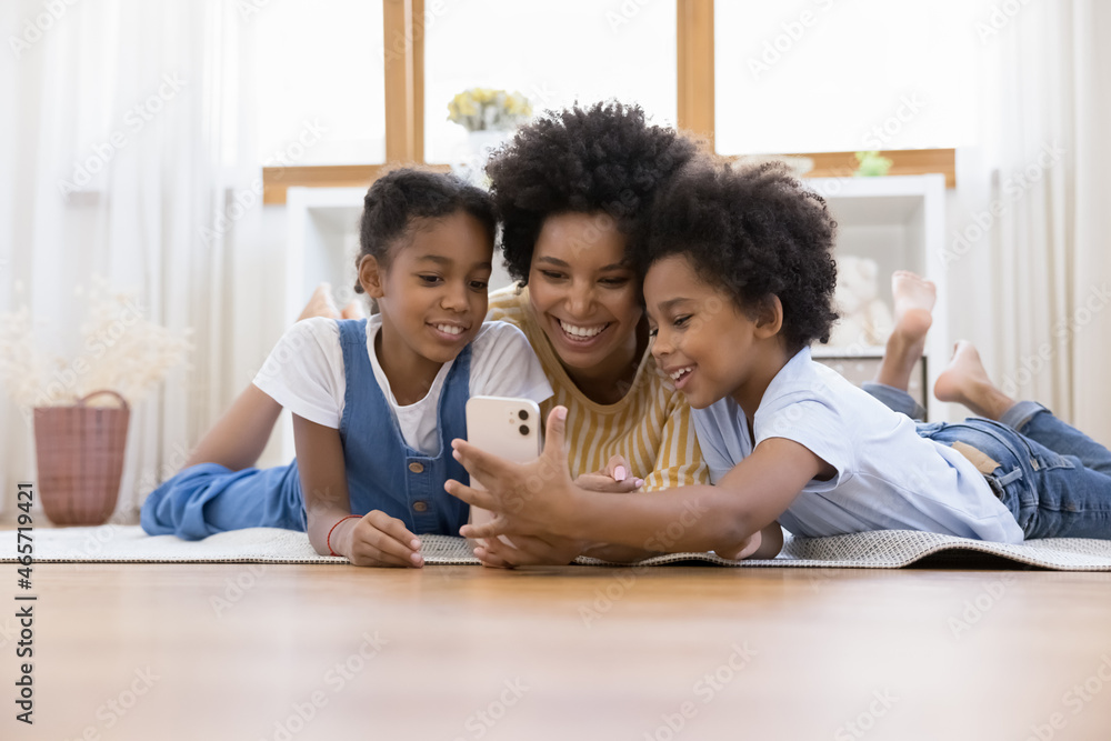 Happy beautiful young african american mother or nanny using cellphone, recording funny video with cute small children siblings, taking photos or playing games, lying on floor carpet together at home. Happy beautiful young african american mother or nanny using cellphone, recording funny video with cute small children siblings, taking photos or playing games, lying on floor carpet together at home.