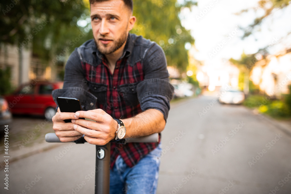Young urban man driving his scooter in the park holding his phone typing a text, wearing checked shirt and a watch, enjoying in the park, looking up to the sky Young urban man driving his scooter in the park holding his phone typing a text, wearing checked shirt and a watch, enjoying in the park, looking up to the sky