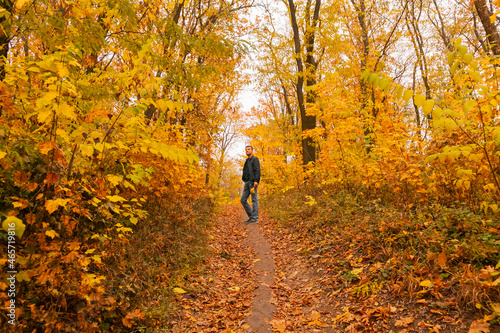 A man is standing on the pathway through autumn forest, among trees and shrubs, a maple leaf in his hands, enjoys autumn and nature © Kathleen
