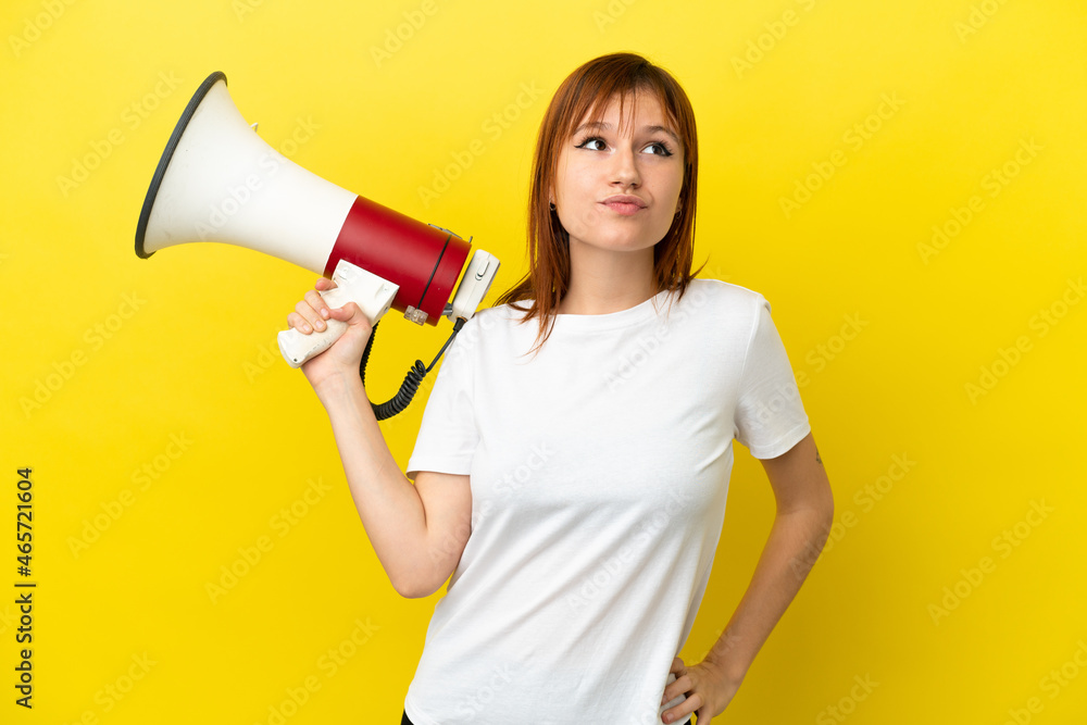 Redhead girl isolated on yellow background holding a megaphone and thinking Redhead girl isolated on yellow background holding a megaphone and thinking