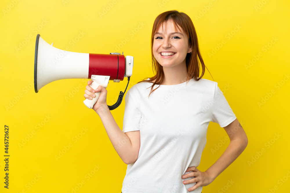 Redhead girl isolated on yellow background holding a megaphone and smiling Redhead girl isolated on yellow background holding a megaphone and smiling