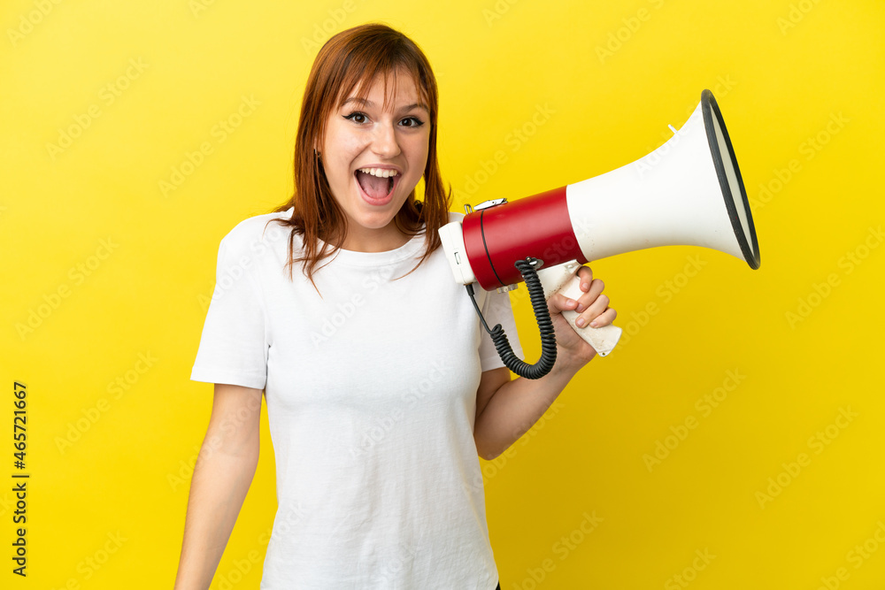 Redhead girl isolated on yellow background holding a megaphone and with surprise expression Redhead girl isolated on yellow background holding a megaphone and with surprise expression