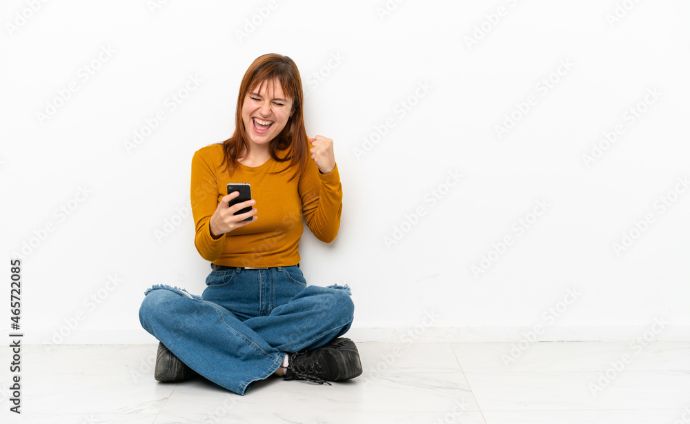 Redhead girl sitting on the floor isolated on white background with phone in victory position Redhead girl sitting on the floor isolated on white background with phone in victory position