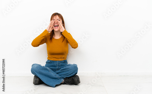 Redhead girl sitting on the floor isolated on white background shouting and announcing something