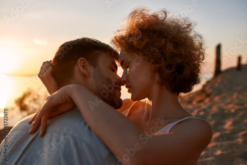 Portrait of passionate young couple closing their eyes before kiss, sitting on the beach at sunset