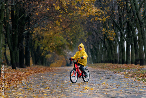 Child rides bicycle in deserted autumn park. Boy in yellow raincoat rides bicycle in rainy autumn park.