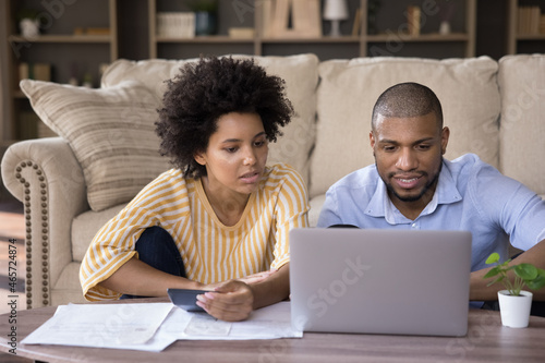 Focused young african american family couple calculating expenses, analyzing paper bills, paying for services in e-banking computer application, managing monthly budget or planning investment at home.