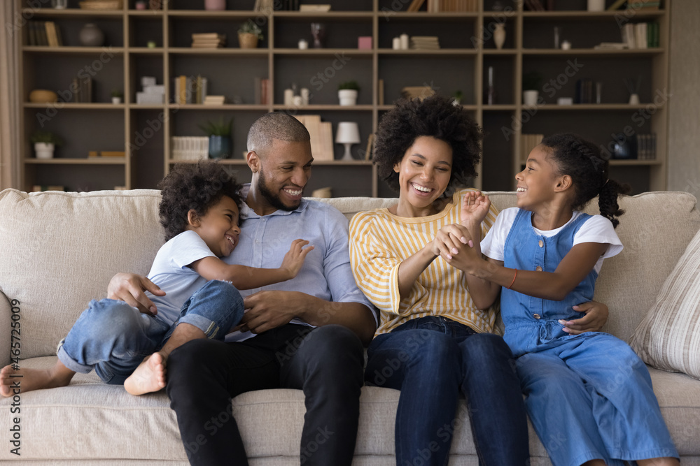 Happy affectionate young african american family couple parents having fun tickling little adorable children son daughter, playing together resting on comfortable sofa, leisure activity concept.
