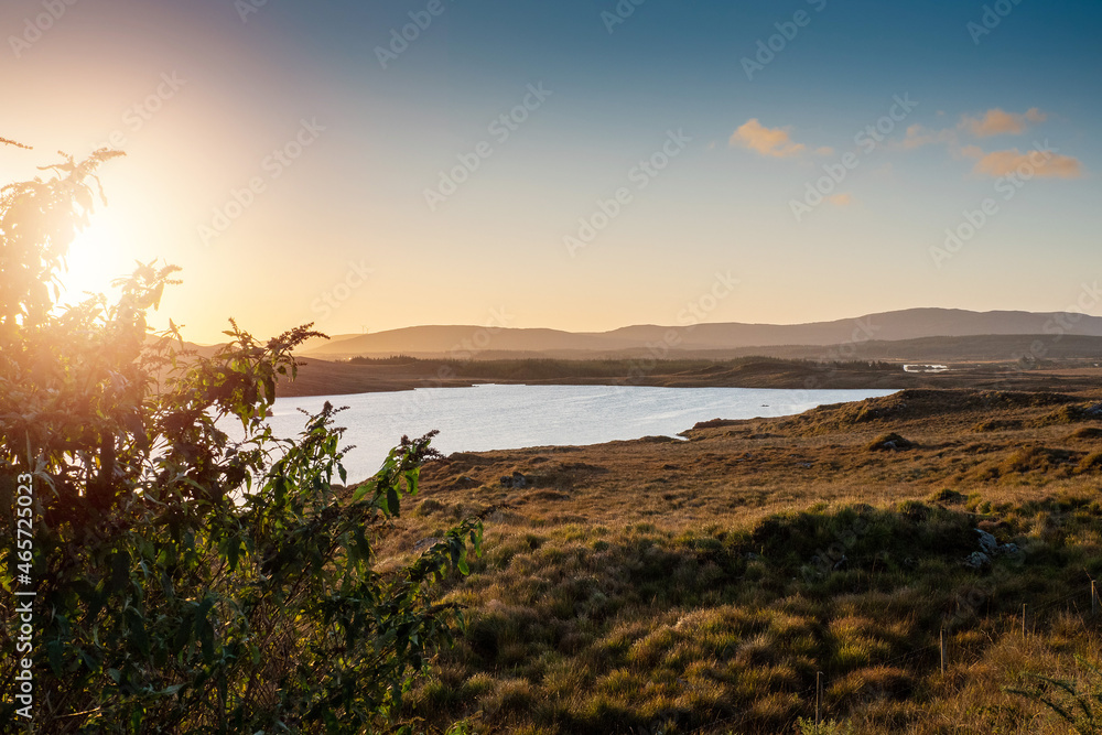 Small lake with blue water and wild fields. Beautiful sun rise scene in Connemara, county Galway, Ireland. Warm and cool tones. Haze over mountains in the background.
