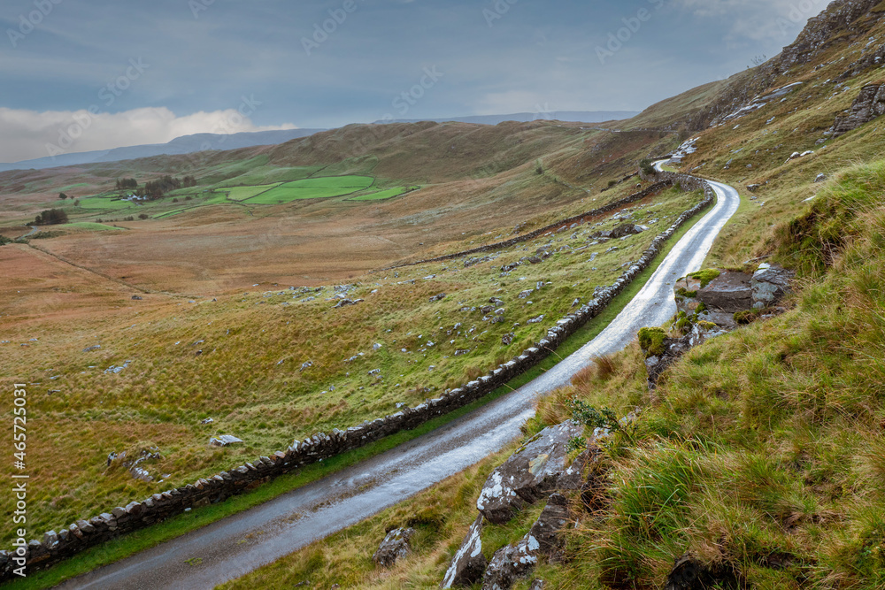 Small narrow country road on a hill by a traditional stone wall with beautiful nature scene. West of Ireland. Irish landscape. Green grass. Fog in the background. Transportation industry.