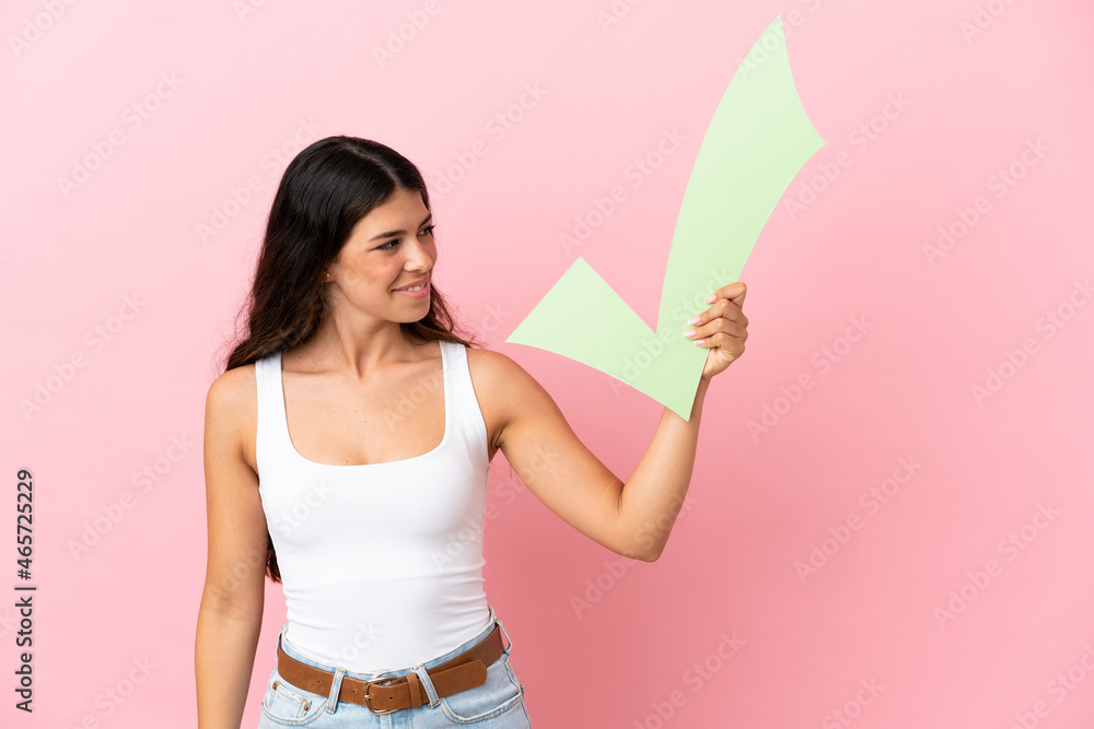 Young caucasian woman isolated on pink background holding a check icon with happy expression Young caucasian woman isolated on pink background holding a check icon with happy expression