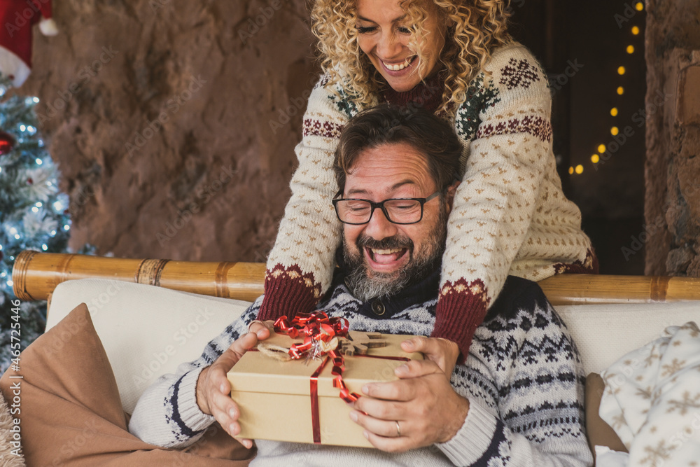 © simona - Christmas eve holidays gif exchange activity with man and woman at home. Female give present at adult male sitting on the sofa. Lady make a surprise tu her husband with box