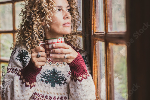 Adult attractive curly blonde woman at home enjoying winter holidays drinking from decorated mug and looking outside wht winwos glass. Female middle age people indoor leisure activity photo