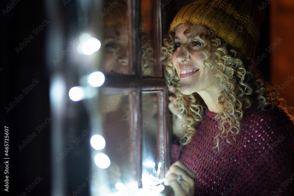 Happy young adult woman look outside the windows at home by night. Female people with curly hair and knit hat enjoy indoor in winter season. Holiday lights and reflection on the glass Happy young adult woman look outside the windows at home by night. Female people with curly hair and knit hat enjoy indoor in winter season. Holiday lights and reflection on the glass
