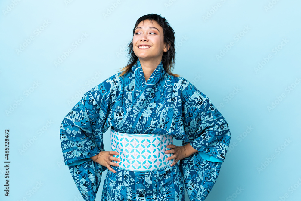 Young woman wearing kimono over isolated blue background posing with arms at hip and smiling Young woman wearing kimono over isolated blue background posing with arms at hip and smiling
