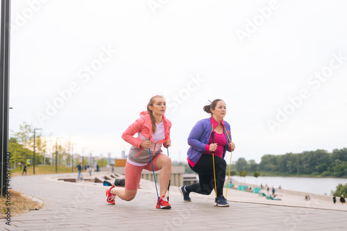 Front view of fat young woman doing squats exercises using fitness tape for weight loss with personal trainer outdoor in summer evening. Overweight female with coach stretching before running.