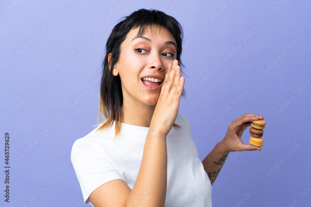 Young Uruguayan woman over isolated purple background holding colorful French macarons and whispering something Young Uruguayan woman over isolated purple background holding colorful French macarons and whispering something