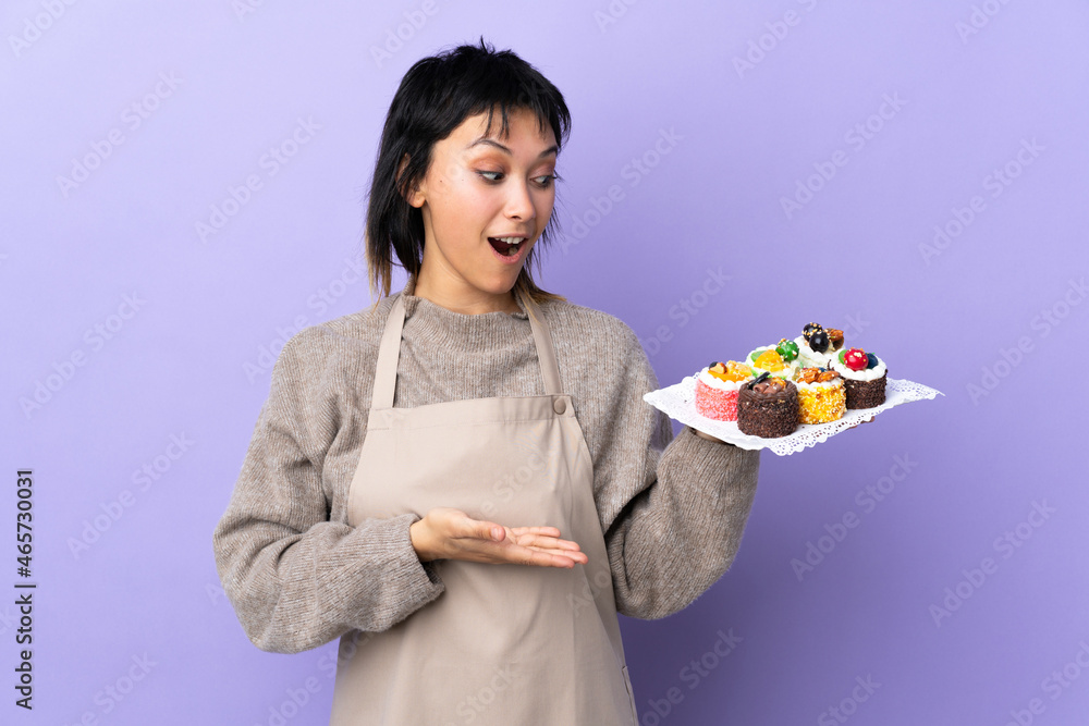 Young Uruguayan woman holding lots of different mini cakes over isolated purple background holding copyspace imaginary on the palm Young Uruguayan woman holding lots of different mini cakes over isolated purple background holding copyspace imaginary on the palm