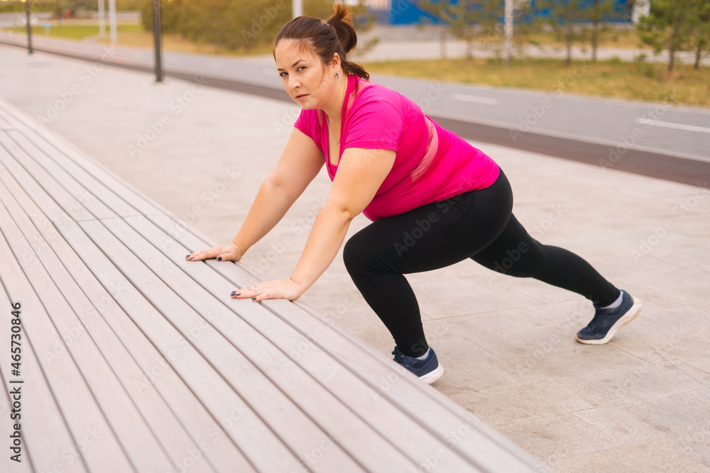 Side view of motivated fat young woman training doing push ups using street bench in city park at summer morning, looking at camera. Fitness female doing bench push ups for strength workout outdoors.