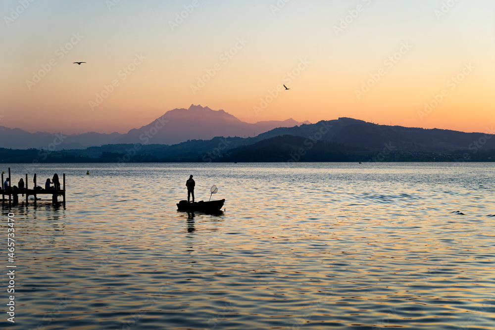 View of Lake Zugersee at sunset, in the Swiss city of Zug. The lake is calm in good weather and the silhouette of a boat with a fisherman.