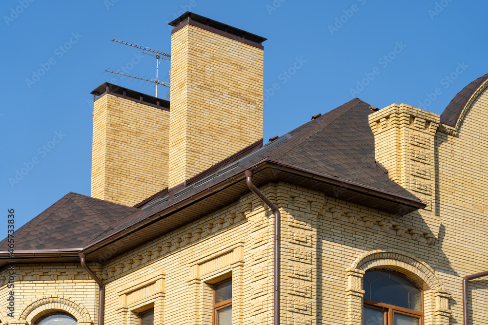 house with drainage system on metal roof. Сhimney pipes is finished externally with natural stone. Roof gutter pipeline system. House rain gutter with holders and downspout pipe house with drainage system on metal roof. Сhimney pipes is finished externally with natural stone. Roof gutter pipeline system. House rain gutter with holders and downspout pipe