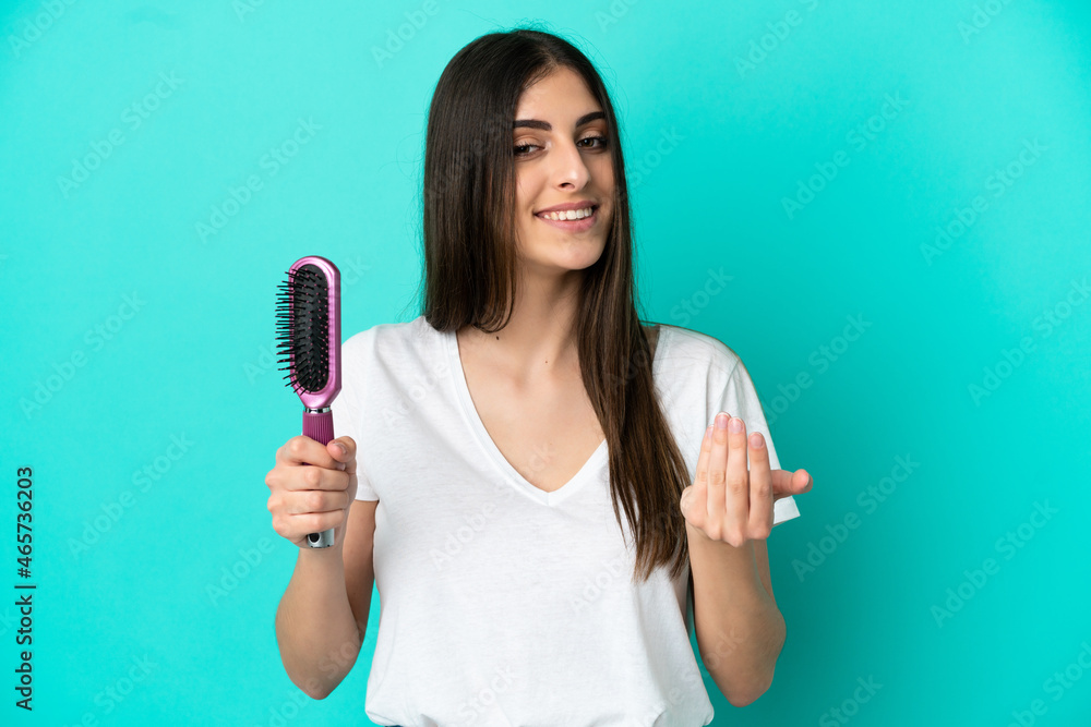 Young caucasian woman with hair comb isolated on blue background inviting to come with hand. Happy that you came Young caucasian woman with hair comb isolated on blue background inviting to come with hand. Happy that you came