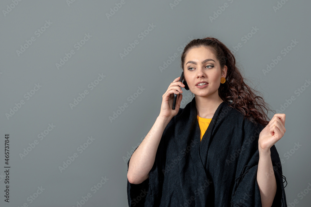 curly-haired emotional young woman in a black cloak talk on mobile phone, isolated on gray curly-haired emotional young woman in a black cloak talk on mobile phone, isolated on gray