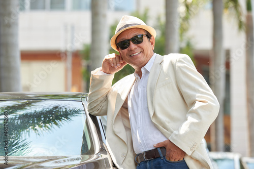 Porteait of positive successful mature Asian man in sunglasses and hat standing at car and leaning on it outdoors photo