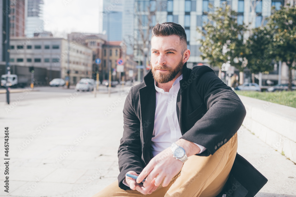 Obraz premium Young adult caucasian bearded man outdoors sitting waiting holding smartphone